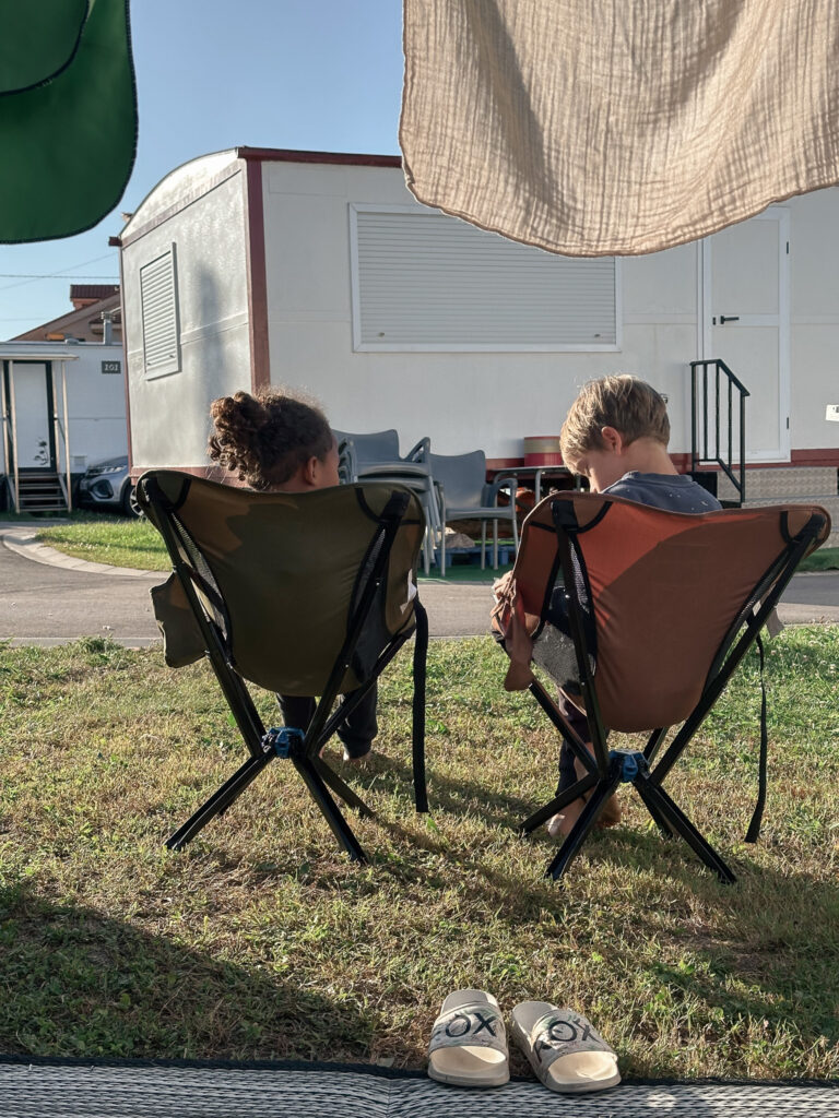 Deux enfants assis de dos dans des fauteuils de camping sur l’herbe, devant un mobil-home