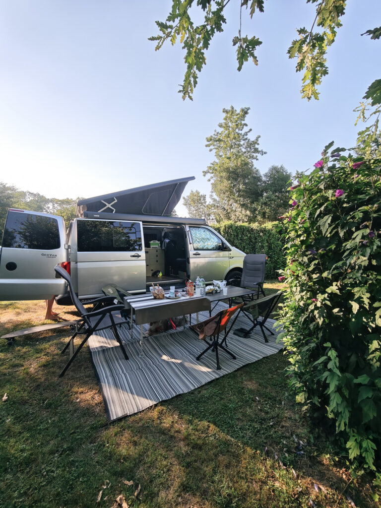 Van avec toit relevé et coin repas installé sur un tapis extérieur, à côté d’une haie fleurie
