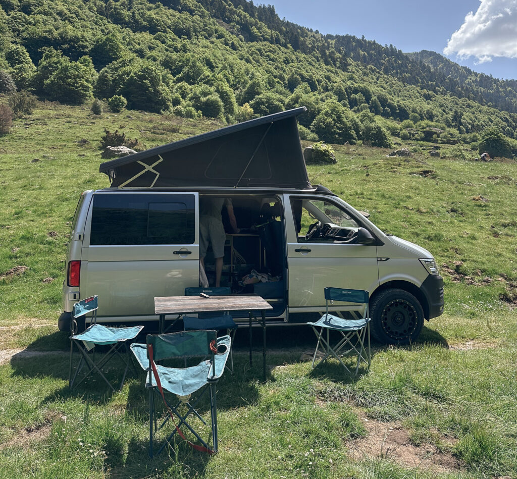 Van avec toit relevé, table et chaises installées sur l’herbe devant une montagne boisée